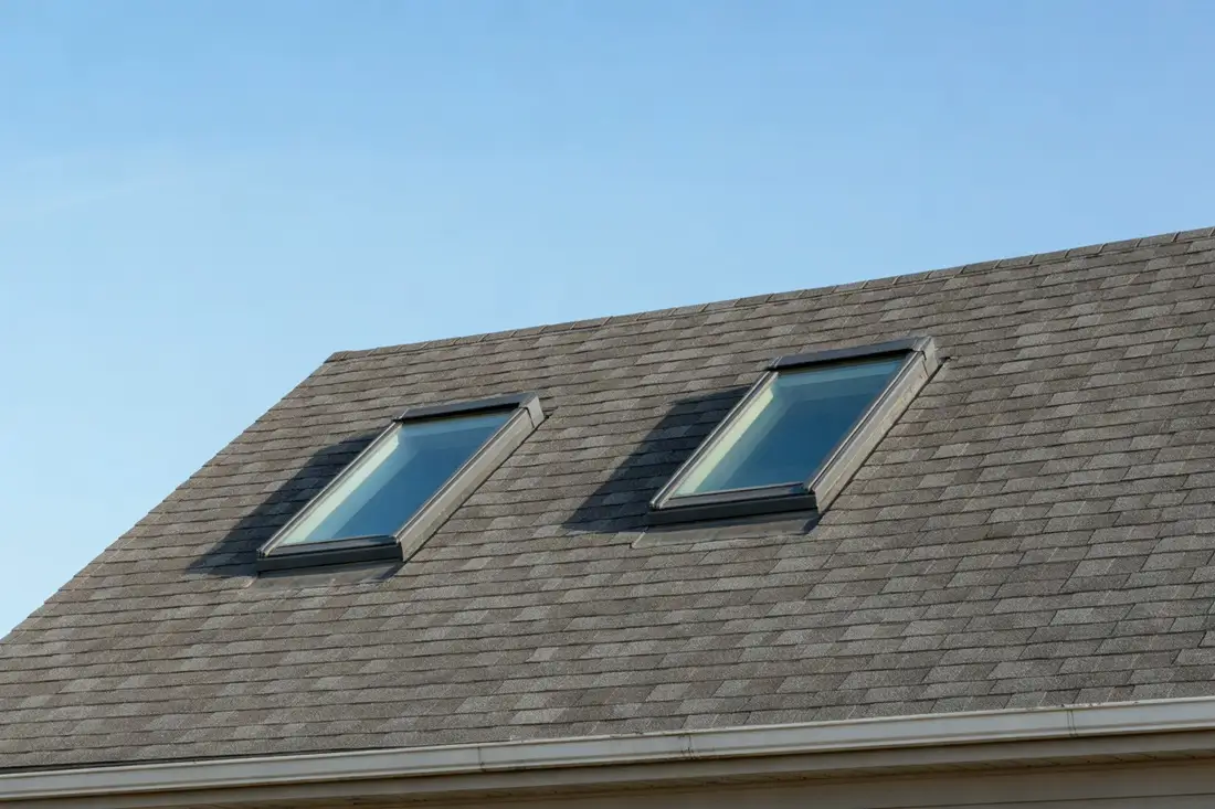 Two glass skylights set flush into a gray asphalt shingle roof under a clear blue sky